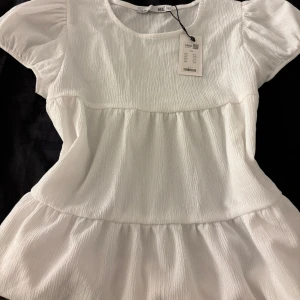 Vit volangblus med puffärm Lager 157 - Superfin vit blus från Lager 157, med lapp på aldrig använd, med volanger och puffiga korta ärmar. Blusen har en lätt rynkad struktur och rund halsringning, vilket ger en söt och luftig känsla. Perfekt för dig som gillar en romantisk och trendig stil.