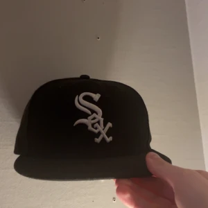 Svart White Sox keps från New Era - Snygg svart keps från New Era med White Sox-logga broderad i vitt framtill. Klassisk flat brim och MLB-logga baktill. Kepsen har en stilren look och passar perfekt för dig som gillar sportiga accessoarer. Storleken är 7/14 eller 57,7cm 
