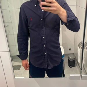 Mörkblå skjorta Ralph Lauren slim fit - Säljer en mörkblå skjorta från Ralph Lauren i slim fit-modell. Skjortan har klassisk krage, vita knappar och den ikoniska röda Polo-loggan broderad på bröstet. Tillverkad i mjuk bomull och har lång ärm. Perfekt för dig som gillar stilrena och tidlösa plagg. OBS tappade en knapp när jag skulle ta bild, men den medföljer så sänker pris därefter. Annars snygg som fan.