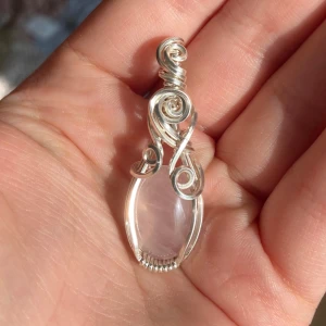 Silverberlock med en rosa rosenkvarts  - Unik berlock med en oval, ljusrosa kristall infattad i silverfärgad tråd som är snirkligt lindad runt stenen. Hängsmycket har coola spiraldetaljer och en elegant, bohemisk vibe. Perfekt för dig som gillar kristaller och vill sticka ut med något personligt.