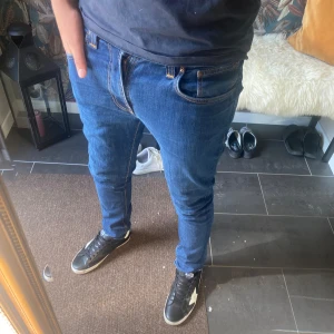 Nudie Jeans - Säljer ett par klassiska blå jeans från Nudie Jeans i modellen slim fit. Jeansen har snygga kontrastsömmar, broderad bakficka och läderpatch i midjan. Tillverkade i mjukt bomullstyg med en smal passform som sitter snyggt längs benen.