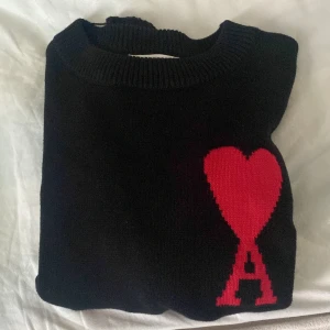 Svart stickad tröja Ami Paris hjärta - Svart stickad tröja från Ami Paris med ikoniskt rött hjärta och bokstaven A på bröstet. Tröjan har rund halsringning och ribbade muddar. Perfekt för dig som gillar stilrena plagg med en twist.