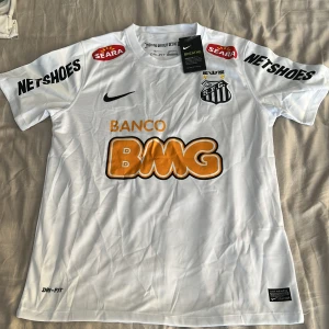 Vit Santos Neymar Jr matchtröja Nike - Santos FC fotbollströja med Neymar Jr och nummer 11 på ryggen. Vit med svarta och orange tryck, flera sponsorer och klubbmärke på bröstet. Tillverkad i Dri-FIT material som andas. Klassisk passform och korta ärmar, perfekt för fans av brasiliansk fotboll.