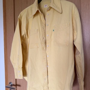 Gul overshirt från Levi's i bomull - Snygg ljusgul overshirt från Levi's i bomull. Skjortan har klassisk krage, två bröstfickor med lock och knappar framtill. Perfekt att bära öppen eller stängd för en chill look. Långärmad modell med knappar vid ärmslut.