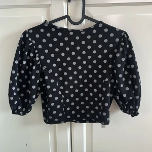 Svart prickig topp från Cameo Rose - Snygg svart topp med silvriga prickar från Cameo Rose. Modellen är croppad med puffiga trekvartsärmar och rund halsringning. Toppens material känns lätt och har en cool struktur, perfekt för att sticka ut lite extra.