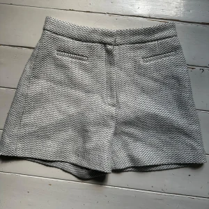 Gråvita strukturerade shorts från Dagmar - Snygga gråvita shorts från Dagmar i storlek 36. De har en vävd, strukturerad yta och klassisk passform med hög midja och diskreta fickor bak. Stängs med dragkedja i sidan. Perfekta för dig som gillar stilrena och trendiga plagg.