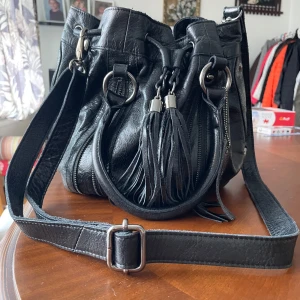 Svart skinnväska med fransar från Enjöd - Genuine leather with long fringes, silver-tone hardware, and zippers. Adjustable strap for comfort. Stylish rounded shape with bold ring details. In great condition — perfect if you love an edgy, standout look! 🖤