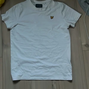 Vit t-shirt från Lyle & Scott Lite större i storleken! - Vit t-shirt från Lyle & Scott med klassisk rund hals och korta ärmar. På bröstet sitter den ikoniska gula örnloggan. T-shirten är i mjuk bomull och har en clean, stilren look som passar perfekt till jeans eller shorts.