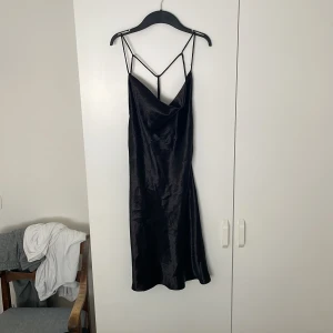 Svart satin klänning Gina Tricot -  svart klänning i satin från Gina Tricot. Klänningen har tunna axelband, draperad urringning och öppen rygg med banddetaljer. 