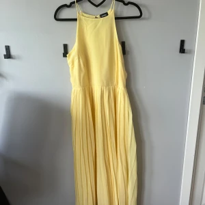Ljusgul långklänning med plisserad kjol - Superfin ljusgul långklänning med plisserad kjol och hög halsringning. Klänningen har ärmlös design och stängs med dragkedja och knapp i ryggen. Perfekt för sommarens alla tillfällen!