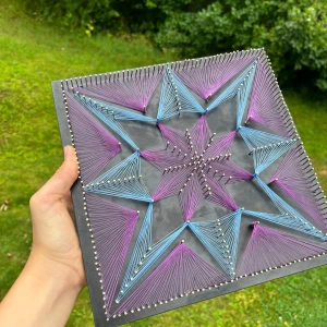 String Art: Stjärnexplosion - Upptäck kreativitet med denna unika string art-tavla! Färgglada trådar i lila och blått bildar en hypnotisk stjärnform på svart bakgrund. Perfekt för dig som älskar DIY, konst och vill ha något coolt och inspirerande på väggen.