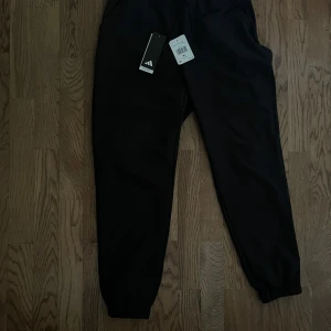 Svarta mjukisbyxor från Adidas M - Svarta mjukisbyxor från Adidas i sportig modell med resår i midjan och benslut. Byxorna har dragsko i midjan och är tillverkade i ett mjukt syntetmaterial. Perfekta för chill eller träning, stilrena och bekväma.