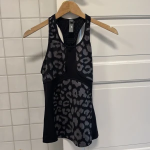 Leopardmönstrad träningstopp Adidas Stella McCartney - Snygg svart träningstopp från Adidas Stella McCartney med grått leopardmönster och sportig racerback. Toppen är ärmlös och tillverkad i ett stretchigt syntetmaterial som andas, perfekt för intensiva träningspass. Logga framtill och cool meshdetalj i ryggen 🥰