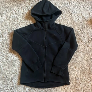 Svart hoodiejacka från Nike - Svart hoodiejacka från Nike med hel dragkedja och huva. Jackan har en sportig look med markerade sömmar och ribbade muddar vid ärmsluten. Perfekt för chill dagar eller när du vill ha en clean streetstyle.