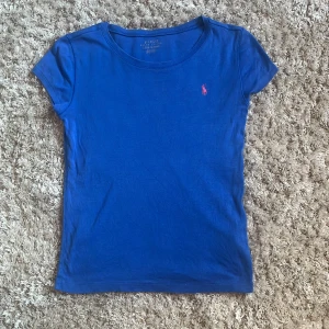 Blå t-shirt från Polo Ralph Lauren - Klassisk blå t-shirt från Polo Ralph Lauren med rund halsringning och korta ärmar. Liten rosa broderad logga på bröstet. Skön och enkel modell i mjuk bomull, perfekt till jeans eller shorts.
