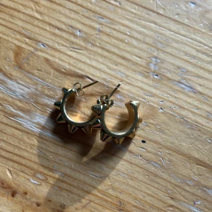 Guldiga hoops med nitar - Coola örhängen i form av hoops med spetsiga nitar runt om. De är i guldig metall och har en edgy vibe som verkligen sticker ut. Perfekta för dig som vill ha något annorlunda och vågat i din smyckessamling.