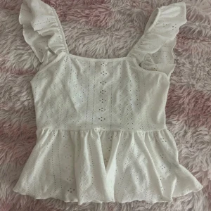 Vit volangblus med broderi och knyt - Supersöt vit blus med volangärmar och broderade detaljer. Toppen har en luftig peplumform, fyrkantig ringning och knyt i ryggen. Perfekt för varma dagar när du vill ha en romantisk och trendig look.