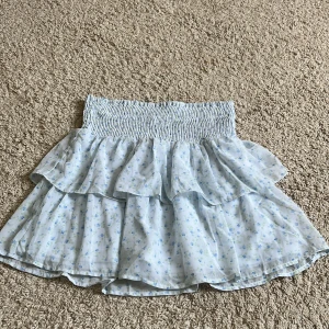 Blommig volangkjol - Supersöt ljusblå kortkjol från Vero Moda med små blå blommor och gröna detaljer. Kjolen har smock i midjan och dubbla volanger som ger en flowy känsla. Perfekt för sommarens alla tillfällen!💘Går att vika ner om man vill ha den lågmidjad😍