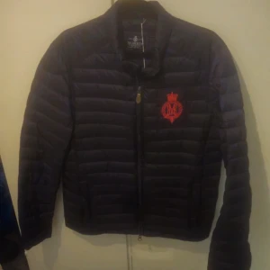 Svart dunjacka från Morris, Small - Svart dunjacka från Morris i storlek Small. Jackan har quiltad design, dragkedja framtill och en snygg röd broderad logga på bröstet. Perfekt för kyliga dagar och riktigt stilren att bära till det mesta. Nypris 2300