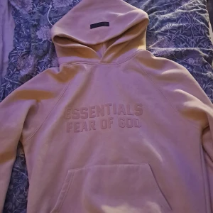 Beige Essentials Fear of God hoodie - Säljer en beige hoodie från Essentials Fear of God med broderad logga framtill. Tröjan har stor huva, känguruficka och en clean, oversized look. Perfekt för dig som gillar streetwear och vill ha något stilrent men ändå trendigt.