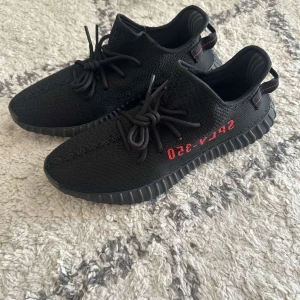 Adidas Yeezy Boost 350 V2 Black - Säljer ett par Adidas Yeezy Boost 350 V2 i svart med röda detaljer och texten SPLY-350 på sidan. Skorna har stickad ovandel, snörning och en tjock, räfflad sula med boost-dämpning. Perfekta för dig som gillar streetwear och vill sticka ut.