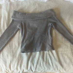 Grå ribbad offshoulder topp Kappahl - Snygg grå ribbad topp från Kappahl med offshoulder-design och långa ärmar. Mjuk och stretchig, perfekt för att lyfta din outfit med en trendig touch. Passar dig som gillar stilrena och enkla plagg med lite edge.