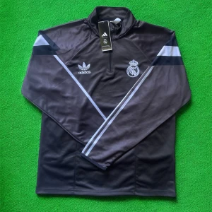 Adidas Real Madrid svart half zip tröja - Svart långärmad Adidas-tröja med Real Madrid-logga och vita detaljer. Tröjan har half zip, högt krage och klassiska Adidas-ränder på ärmarna. Tillverkad i polyester, perfekt för fotboll eller träning. Snygg sportig look med broderade loggor framtill.