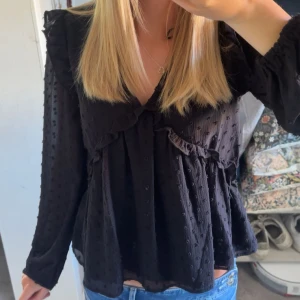 Svart blus med volang från Gina Tricot - Supersöt svart blus från Gina Tricot med v-ringning och volangdetaljer längs bröstet och axlarna. Tunt prickigt material. Använd fåtal gånger💗 