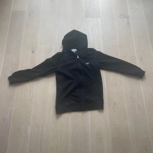 Svart hoodiejacka från Lacoste us  M - Svart hoodiejacka från Lacoste med dragkedja framtill och klassisk krokodillogga på bröstet. Jackan har huva, långa ärmar och två fickor framtill. Perfekt för en chill och sportig stil.