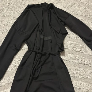 Svart omlott kappa med bälte - Svart långärmad kappa i omlottmodell med bälte i midjan. Jackan har en stilren och enkel design med mjukt, lätt material som ger ett snyggt fall. Perfekt för dig som gillar minimalistisk stil och vill ha något som funkar till många outfits.