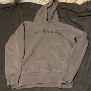 Grå hoodie från Sail Racing - Säljer en stilren grå hoodie från Sail Racing i storlek S. Tröjan har en klassisk huva med snörning, stor magficka och svart logga framtill. Perfekt för dig som gillar en enkel och clean look. Mjuk insida och ribbade muddar för extra komfort.