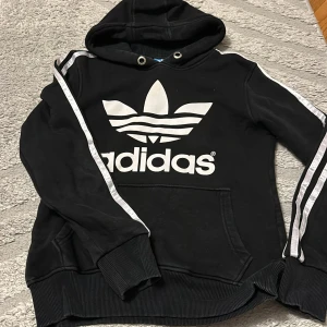 Svart Adidas hoodie med vita ränder - Svart hoodie från Adidas med stor vit logga och text på bröstet. Klassiska vita ränder längs ärmarna och huva med snörhål. Mjuk bomullskänsla och känguruficka framtill. Perfekt för en sportig och avslappnad stil.