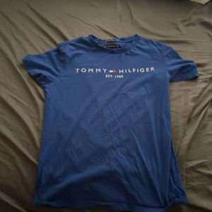 Blå Tommy Hilfiger t-shirt - Snygg blå t-shirt från Tommy Hilfiger med klassisk logga och texten 'EST. 1985' på bröstet. T-shirten har rund halsringning och korta ärmar, perfekt för en chill och stilren look.