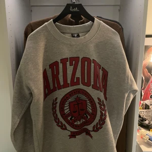 Grå Arizona college-tröja  - Snygg grå sweatshirt från FSBN i relaxed fit med stort rött Arizona State University-tryck framtill. Klassisk rund halsringning och långa ärmar. Perfekt för dig som gillar college-stil och vill ha en bekväm och trendig tröja. Storlek S. Aldrig använd