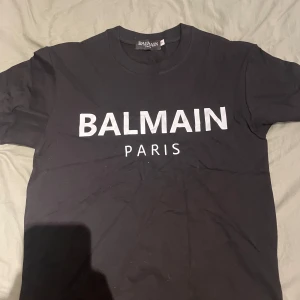 Svart Balmain Paris t-shirt - Svart t-shirt från Balmain Paris med stor vit logga framtill. Klassisk rund hals och korta ärmar. Tillverkad i mjuk bomull som känns skön mot huden. Perfekt för dig som gillar stilrena och trendiga plagg med exklusiv känsla.