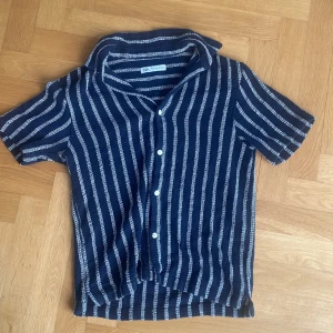 Randig blå skjorta från Zara - Kortärmad skjorta från Zara i marinblått med vita vertikala ränder. Skjortan har klassisk krage, knäppning framtill med vita knappar och är tillverkad i ett luftigt stickat tyg som passar perfekt till varma dagar. Snygg och avslappnad stil. Lägger sig snyggt över axlar och överkroppen 