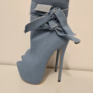 Blå denim boots med platå och rosett - Unika boots i ljusblå denim med hög platå och smal klack. Skorna har öppen tå, grov rosettdetalj och råa kanter för en edgy look. Perfekta för dig som vill sticka ut med statement-skor i jeansmaterial.