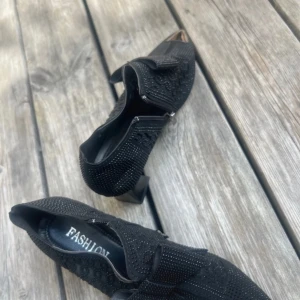 Svarta spetsiga skor med rosett och nitar - Unika svarta skor med spetsig tå och coola silvriga metalldetaljer framtill. Skorna har en stor rosett på ovansidan, små nitar över hela ytan och ett mönstrat mesh-tyg på sidorna. Liten klack och platt sula ger en edgy vibe till outfiten.