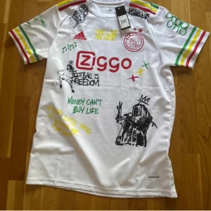 Ajax x Bob Marley Adidas fotbollströja - Unik vit Ajax fotbollströja från Adidas med färgglada detaljer i rött, gult och grönt på ärmarna. Tryck med Bob Marley, graffiti-inspirerade texter och Ajax-logga.  perfekt för match eller träning.