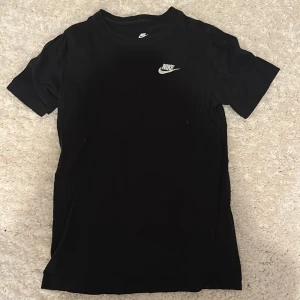 Svart Nike t-shirt med logga - Svart t-shirt från Nike med klassisk vit logga på bröstet. Enkel och stilren design med rund hals och korta ärmar. Perfekt för dig som gillar sportiga och cleana plagg. Tillverkad i mjuk bomull för skön känsla hela dagen. Barn storlek 147-158