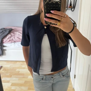 Mörkblå kortarmad kofta från Gina Tricot - Kort, mörkblå kofta från Gina Tricot i storlek XS. Jackan har halvlånga ärmar, rund halsringning och stängs med en dekorativ knapp upptill. Perfekt att slänga över en topp för en clean och stilren look. Använd en gång. Hör av er vid frågor💕