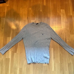 Grå kabelstickad tröja från Ralph Lauren - Snygg grå kabelstickad tröja från Ralph Lauren med klassisk rund halsringning och diskret logga broderad på bröstet.