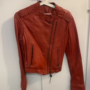 Röd skinnjacka med asymmetrisk dragkedja - Snygg röd skinnjacka med coola detaljer och asymmetrisk dragkedja framtill. Jackan har två fickor med dragkedja, markerade axlar och lång ärm. Perfekt för dig som vill sticka ut och addera lite edge till din outfit. Vintage Levis 