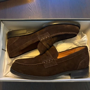 Bruna mockaloafers från Vagabond - Snygga loafers i mörkbrun mocka från Vagabond. Använda 1 gång. Skorna har klassisk design med dekorativ rem över vristen, rund tå och klack. Perfekta för en stilren look och passar till både jeans och kostym.