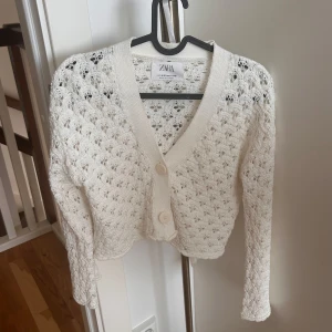 Vit stickad kofta från Zara - Superfin vit stickad kofta från Zara med snyggt hålmönster och två stora knappar framtill. Koftan har v-ringning, lång ärm och är croppad i modellen. Perfekt att slänga över en topp för en chill och trendig look.