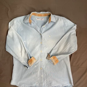 Burberry skjorta storlek M  - Säljer en ljusblå skjorta från Burberrys of London i 100% bomull. Skjortan har klassisk krage, långa ärmar och knappar framtill. Insidan av krage och manschetter har det ikoniska rutiga Burberry-mönstret. Perfekt för dig som gillar klassisk stil med en twist. Den är i okej stil då den är mycket sliten( se bild 4-5). Därför blir den billigare ⭐️⭐️