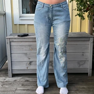 Ljusblå utsvängda jeans med broderi  - Säljer ett par ljusblå jeans med snyggt broderi och applikationer i grönt och beige på ena benet. Jeansen har normal midja, utsvängda som jag sytt själv och klassisk femficksmodell. Perfekta för dig som vill ha något unikt och trendigt i garderoben. Buda på!!