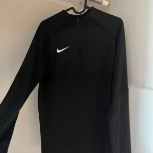 Svart Nike Dri-Fit träningströja - Svart långärmad träningströja från Nike med Dri-Fit-teknologi och slim fit-passform. Tröjan har halv dragkedja framtill och diskret mönster på ärmarna. Perfekt för dig som vill ha en sportig look och funktionellt material.