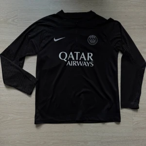 Svart PSG tracksuit storlek M. - Säljer en svart tracksuit  från Nike med Paris Saint-Germain-logga och Qatar Airways-tryck på bröstet. Tröjan är i funktionsmaterial och har diskreta mönster på ärmarna. Perfekt för träning eller när du vill representera PSG. 