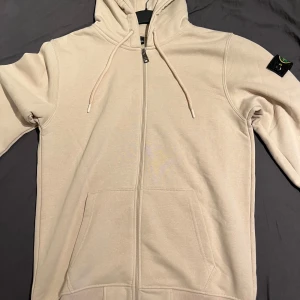 Beige hoodie från Stone Island Storlek L - Snygg beige hoodie från Stone Island i storlek L. Klassisk modell med dragkedja, huva med snörning och känguruficka. Ikonisk logga på vänster ärm. Perfekt för dig som gillar streetwear och vill ha en clean look.
